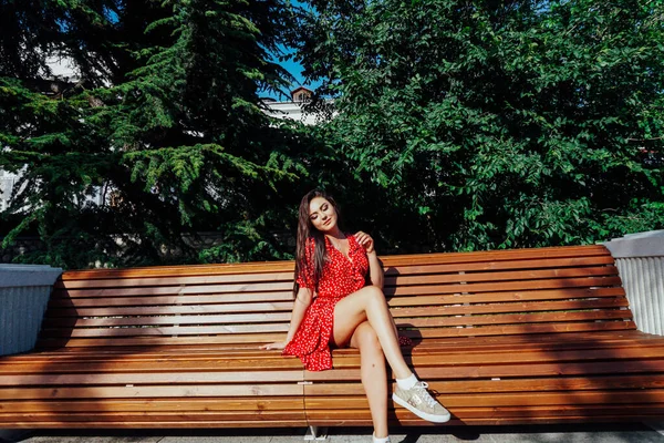 Schöne brünette Frau im roten Kleid sitzt auf einer Bank auf der Straße — Stockfoto