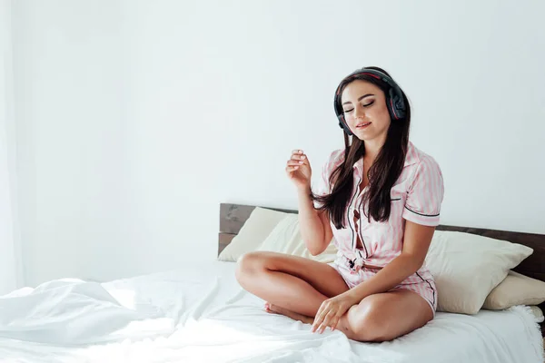 Krásná brunetka žena v růžovém pyžamu poslouchá hudbu ve sluchátkách na posteli — Stock fotografie