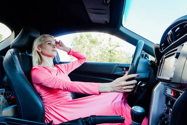 Piękna blondynka kierowca w różowej sukience za kierownicą samochodu — Zdjęcie stockowe