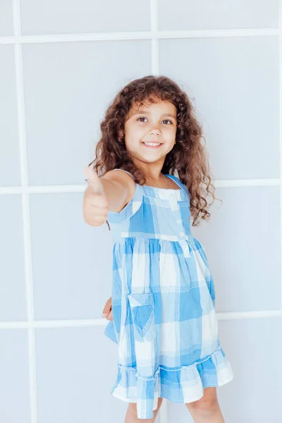 Bella piccola ragazza riccia in abito blu mostra pollice in su — Foto Stock