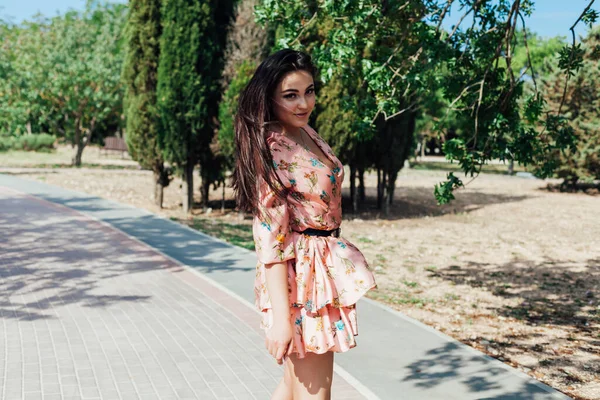 Çiçekli elbiseli güzel, şık esmer kadın parkta yürüyor. — Stok fotoğraf