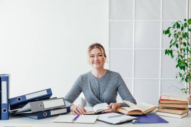 İş elbiseli bir kadın iş yerinde kitap ve belgelerle dolu bir masada.