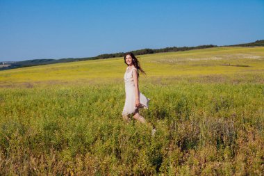 Açık renk elbiseli güzel bir kadın elinde çiçeklerle sahada yürüyor.