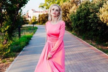 Pembe elbiseli güzel sarışın kadın gün batımında parkta yürüyor.