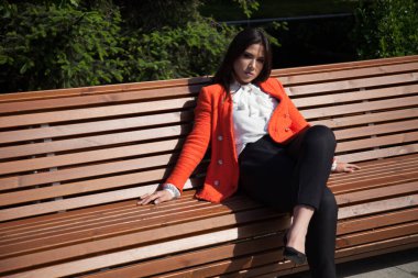 Kırmızı ceketli güzel esmer kadın parkta bir bankta oturuyor.