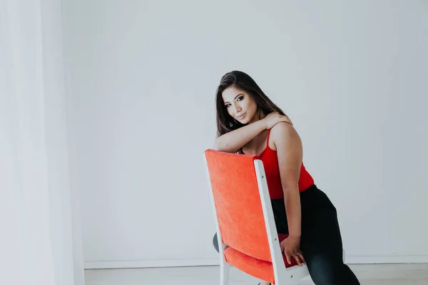 Beautiful brunette woman sits on a red chair in a white room — Stock Photo, Image