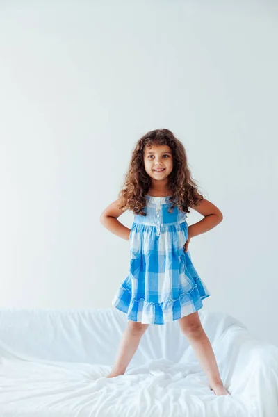 Bella piccola ragazza riccia in un abito blu divertente su un divano bianco — Foto Stock