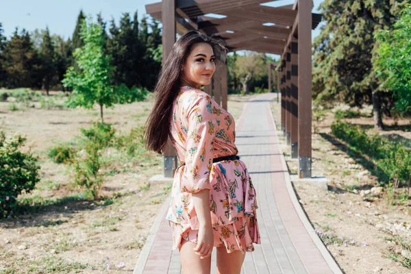 Mulher morena bonita em um vestido com flores caminha no parque sozinha — Fotografia de Stock