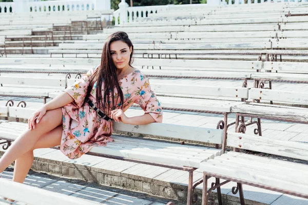 Schöne brünette Frau in einem Kleid mit Blumen sitzt allein auf einer Bank — Stockfoto