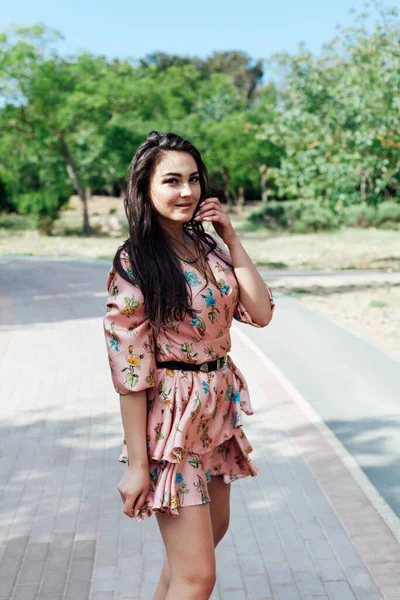 Beautiful brunette woman in a dress with flowers walks in the park alone — Stock Photo, Image