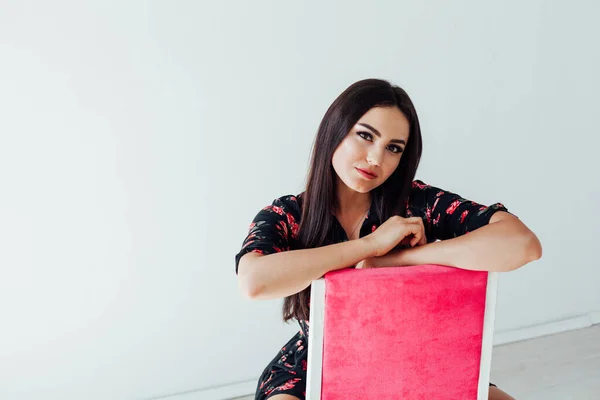 Mooie brunette vrouw in zwarte jurk met bloemen zit op een roze stoel — Stockfoto