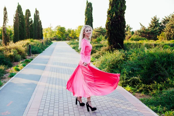 Όμορφη ξανθιά γυναίκα σε ροζ μοντέρνο φόρεμα σε μια βόλτα στο πάρκο στο ηλιοβασίλεμα — Φωτογραφία Αρχείου