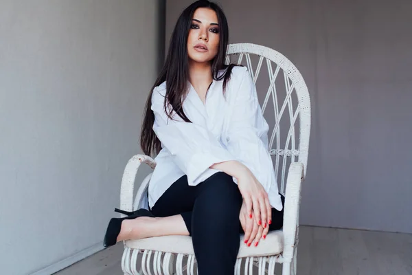 Hermosa mujer morena de moda en una camisa blanca se sienta en una silla — Foto de Stock