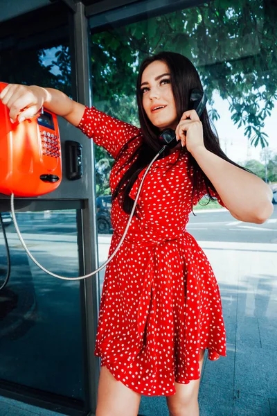 Schöne brünette Frau im roten Kleid spricht in einer Telefonzelle — Stockfoto