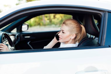 Güzel sarışın şoför, arabanın direksiyonunda akıllı telefondan konuşuyor.