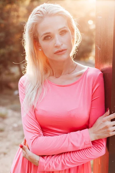 Красивая блондинка в розовом платье гуляет по парку на закате — стоковое фото