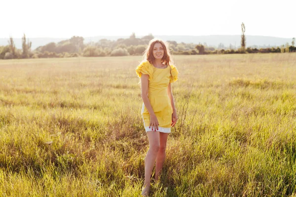 Beautiful woman in yellow dress walks on the field at sunset — Stock Photo, Image