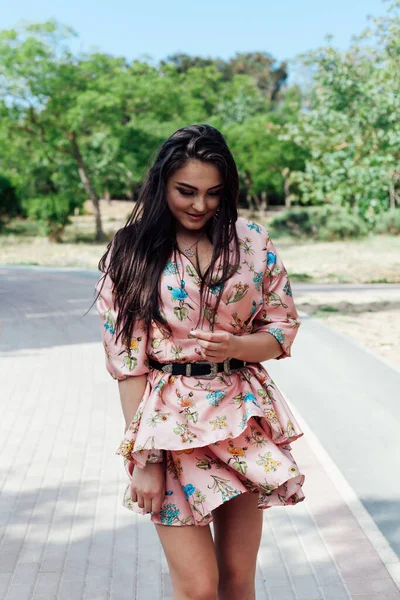 Schöne modische brünette Frau in einem Kleid mit Blumen geht im Park auf der Straße — Stockfoto