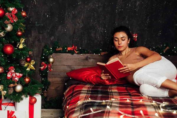Красивая женщина читает книгу в постели у елки с новогодними подарками — стоковое фото