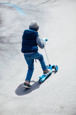 Küçük çocuk parktaki yollarda scooter sürüyor.