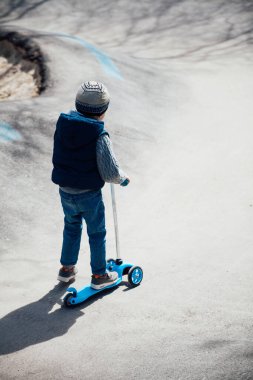 Küçük çocuk parktaki yollarda scooter sürüyor.