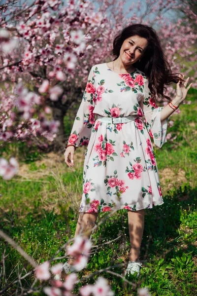 Mulher morena bonita em um vestido com flores caminha pelo jardim florido na primavera — Fotografia de Stock