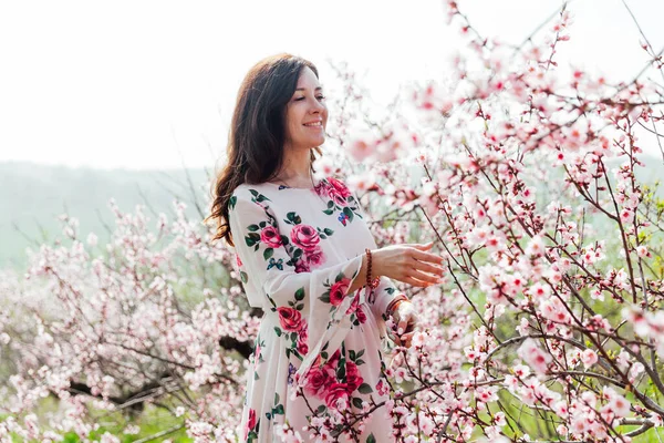 Mulher morena bonita em um vestido com flores caminha pelo jardim florido na primavera — Fotografia de Stock