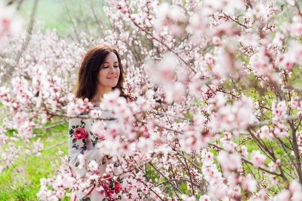 Çiçekli elbiseli güzel esmer kadın baharda çiçek açan bahçede yürüyor. — Stok fotoğraf