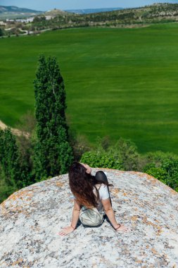 Esmer kadın uçurumun kenarında oturmuş yolculuğun doğasına bakıyor.