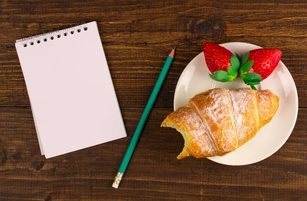 Notebook, croissant och jordgubbar på ett träbord. bästa utsikt. — Stockfoto