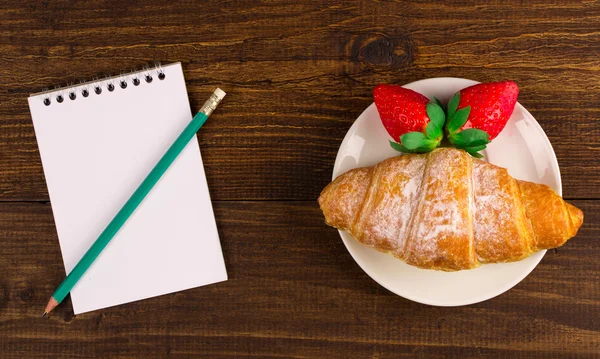 Notebook, croissant och jordgubbar på ett träbord. bästa utsikt. — Stockfoto
