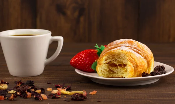 Eine Tasse schwarzen Tee mit Trockenfrüchten, Croissant mit Erdbeere und Holztischhintergrund — Stockfoto