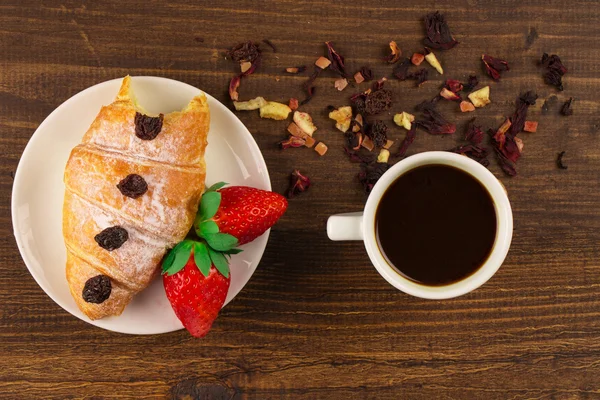 Una taza de té negro con frutas secas, croissant con fresa y fondo de mesa de madera. Arriba wiev . —  Fotos de Stock