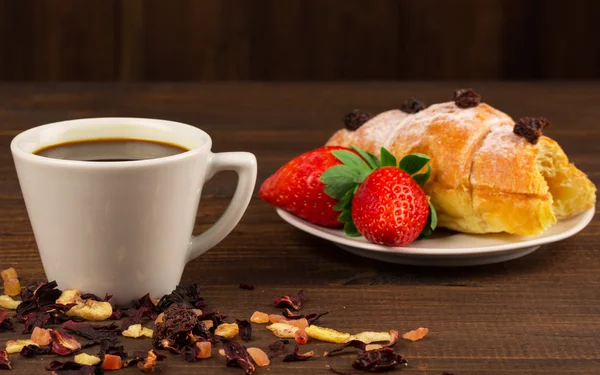 Eine Tasse schwarzen Tee mit Trockenfrüchten, Croissant mit Erdbeere und Holztischhintergrund — Stockfoto