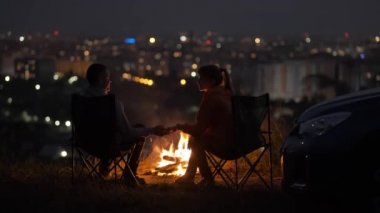 Birbirine aşık genç bir çift gece şenlik ateşinin yanındaki sandalyelerde oturup sıcak romantik bir akşamın tadını çıkarıyor..