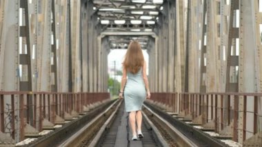 Uzun saçlı, yaz elbisesi giymiş, tren raylarında yürüyen zayıf bir kadının arka görüntüsü..