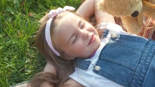 夏天的时候 紧闭双眼的漂亮小女孩躺在绿草上 毛毯上的泰迪熊玩具正在打盹 — 图库视频影像