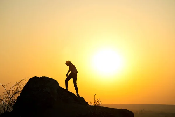 Silhouette of a woman hiker climbing up a big stone at sunset in mountains. Female tourist on high rock in evening nature. Tourism, traveling and healthy lifestyle concept.