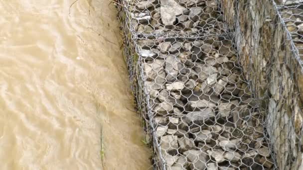 二つの石と金属網で作られた三角形の浸食と洪水保護バリア 大雨による洪水期の濁った川からの堤防の強化 — ストック動画