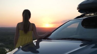 Yazlık elbiseli mutlu genç kız arabasının yanında duruyor ve günbatımının tadını çıkarıyor. Seyahat ve tatil kavramı.