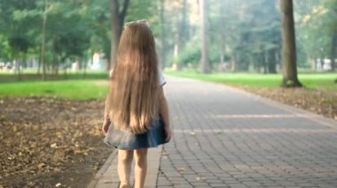 Yeşil parkta tek başına yürüyen yazlık elbiseli küçük bir kızın arka görüntüsü..