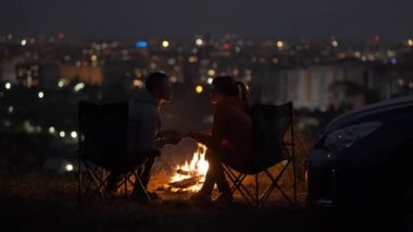 Geceleri yanan sarı kamp ateşinin yanında kamp sandalyelerinde oturan mutlu çift..