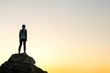 Gün batımında dağlarda büyük bir taşın üzerinde tek başına duran bir kadın yürüyüşçünün silueti. Akşam vakti yüksek kayalıklarda gezen kadın turist. Turizm, seyahat ve sağlıklı yaşam tarzı konsepti.