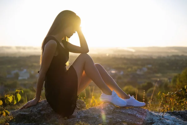 夏天的傍晚 穿着黑色短裙和白色运动鞋的年轻优雅女人坐在岩石上放松自己 时尚的女士享受大自然温暖的落日 — 图库照片