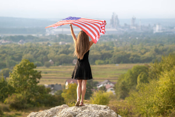 Young happy woman with long hair raising up waving on wind american national flag in her hands standing on high rocky hill enjoying warm summer day.