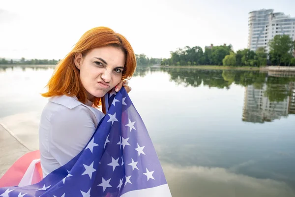 Portrait of sad red haired girl with USA national flag on her shoulders. Young woman celebrating United States independence day.