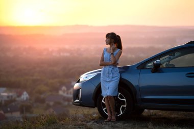 Mavi elbiseli mutlu genç kadın sürücü sıcak yaz akşamının tadını çıkarıyor arabasının yanında duruyor. Seyahat ve tatil kavramı.