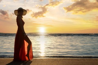 Uzun kırmızı elbise ve hasır şapka giyen genç bir kadın deniz kıyısındaki kumsalda duruyor yaz sabahının erken saatlerinde doğan güneşin tadını çıkarıyor..