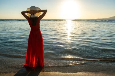 Uzun kırmızı elbise ve hasır şapka giyen genç bir kadın sahildeki deniz suyunda duruyor yaz sabahının erken saatlerinde doğan güneşin tadını çıkarıyor..