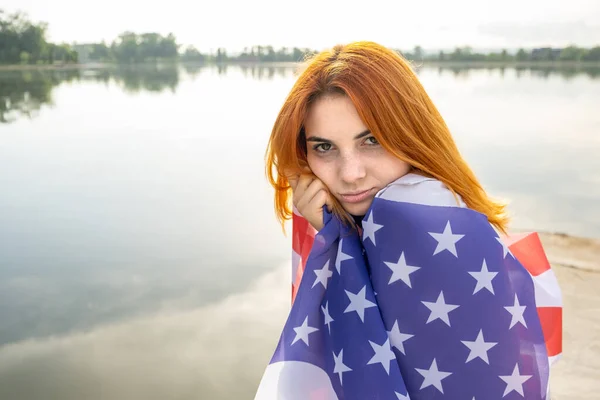 Portrait of sad red haired girl with USA national flag on her shoulders. Young woman celebrating United States independence day.